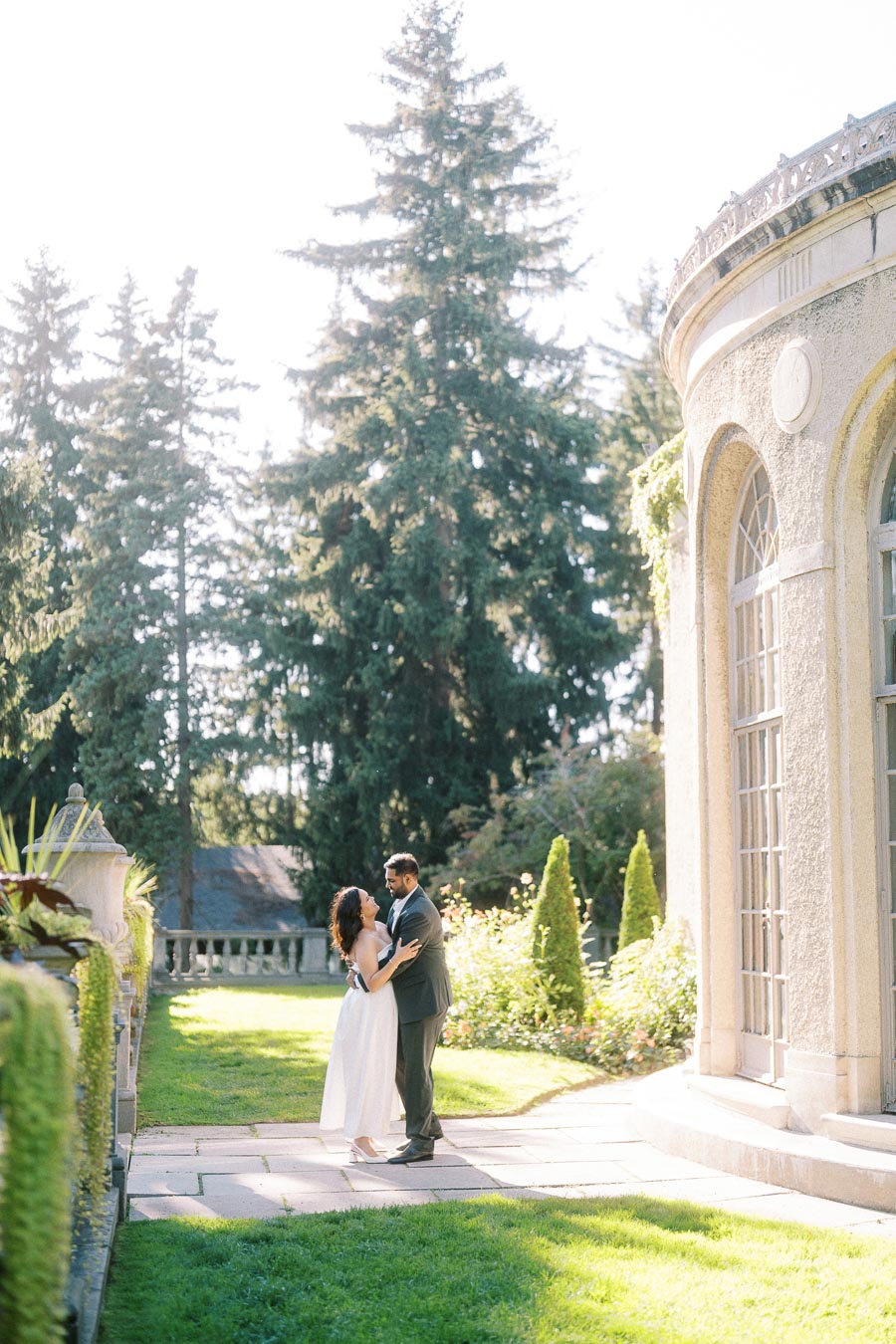 A couple in elegant attire embrace near an ornate building in a sunlit garden, surrounded by lush greenery and tall pine trees.