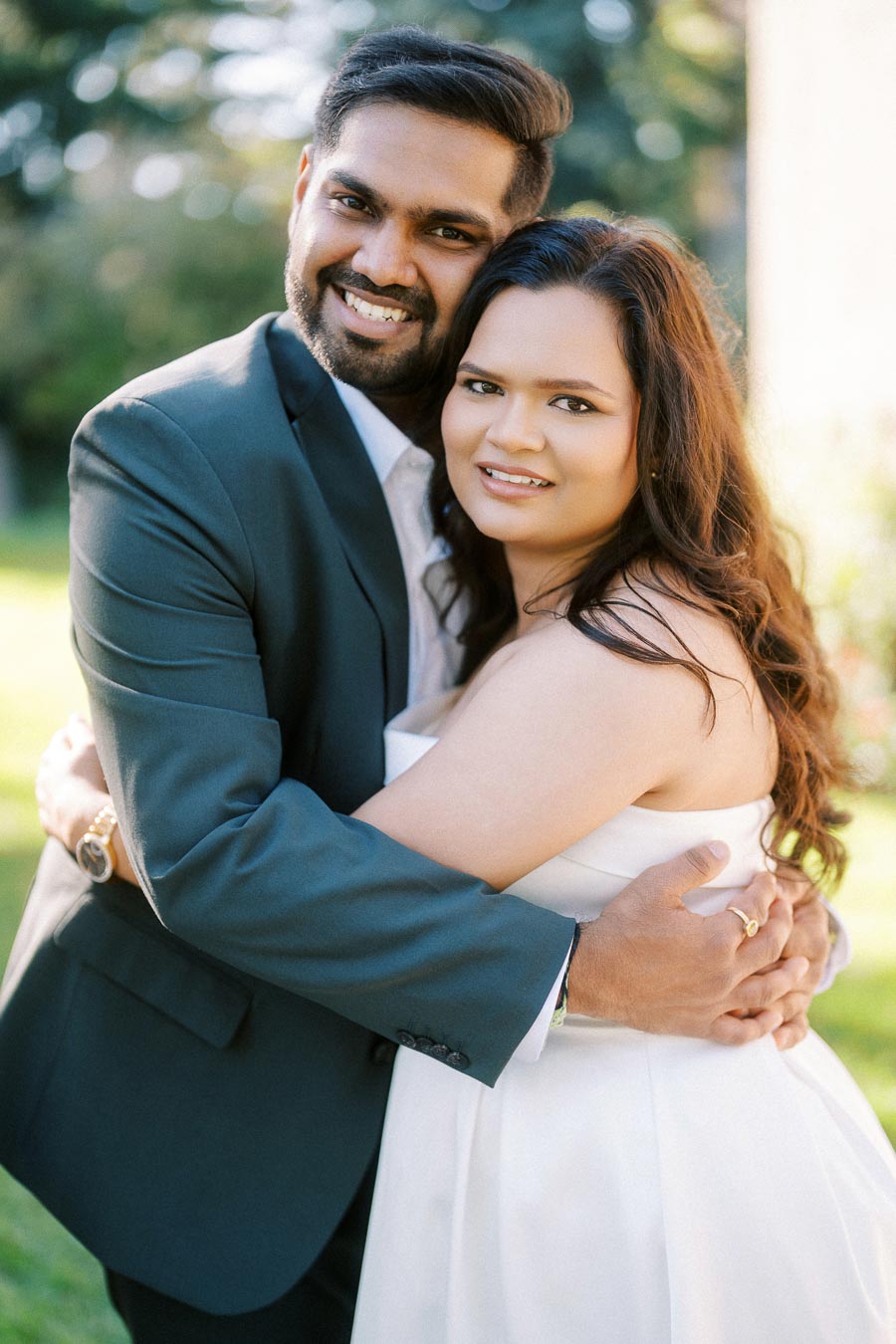 A couple embracing outdoors in a sunlit garden, both smiling warmly, with the man wearing a dark suit and the woman in a white dress.