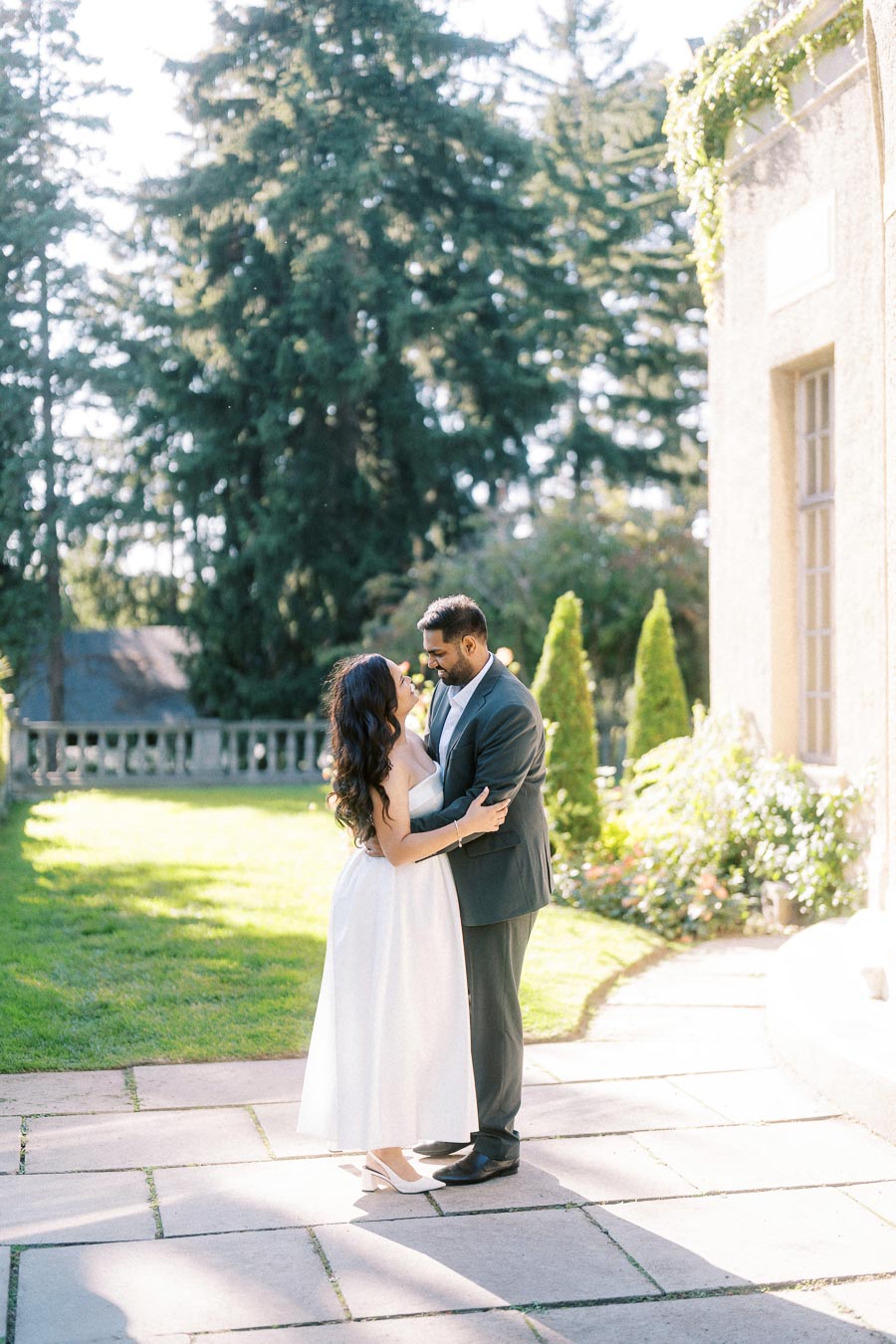 A couple embracing in a sunlit garden, surrounded by trees and greenery, next to a historic building with vine-covered walls.