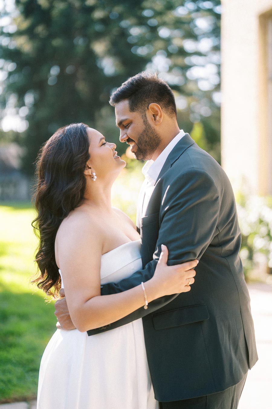 A couple embraces outdoors, smiling at each other in a tender moment, with lush greenery and sunlight in the background.