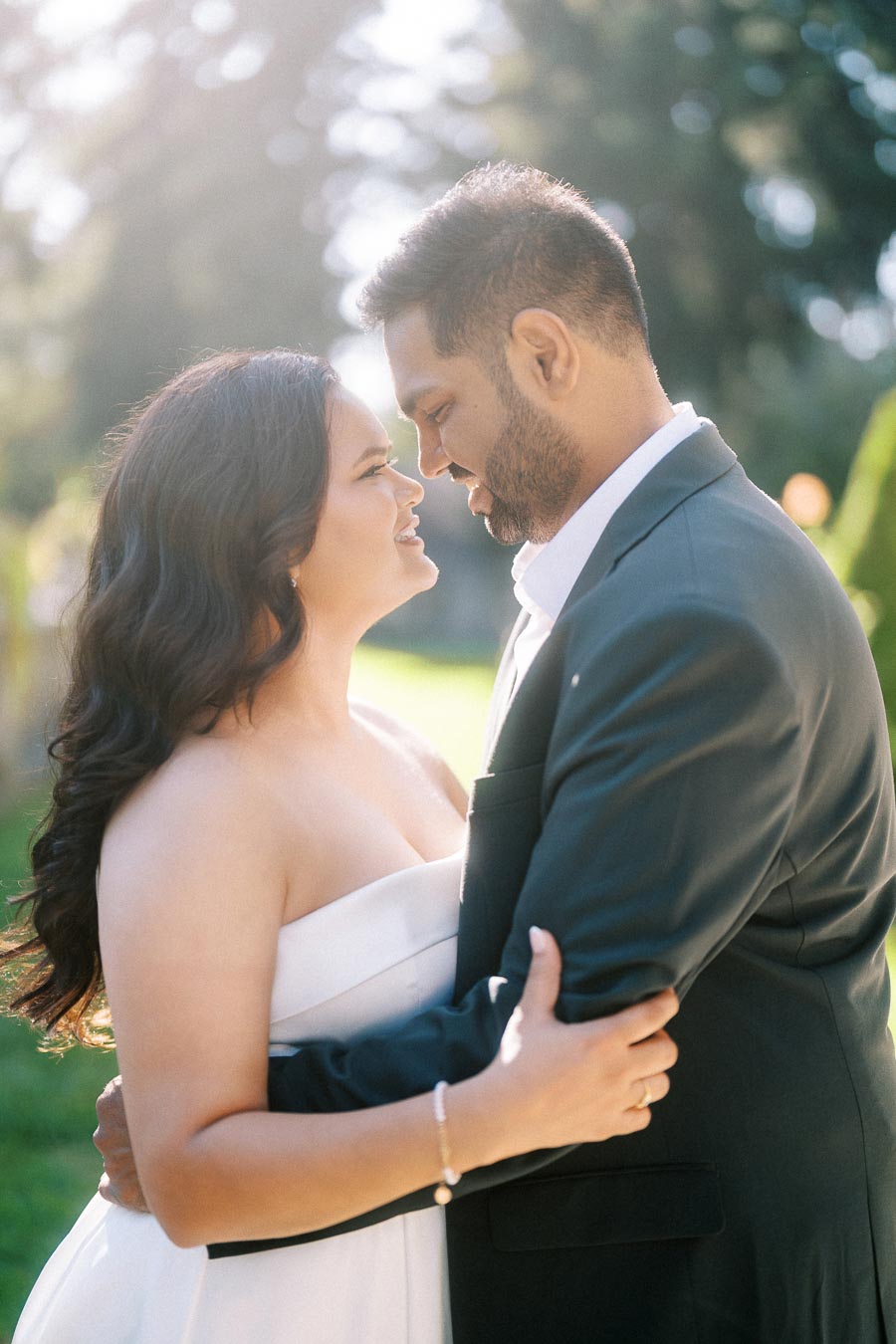 A couple embracing lovingly outdoors in bright sunlight, wearing formal attire, with a blurred natural background.