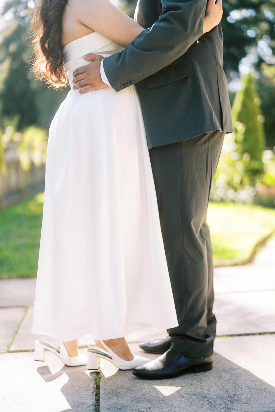 Elegant couple embracing outdoors, bride in white dress and groom in dark suit, with sunlight in the background.