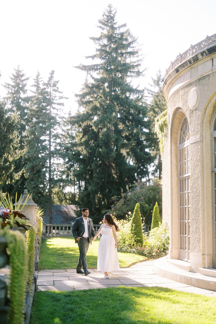 A couple walking hand in hand on a sunlit garden path next to an elegant building, surrounded by lush greenery and tall trees, creating a romantic outdoor scene.
