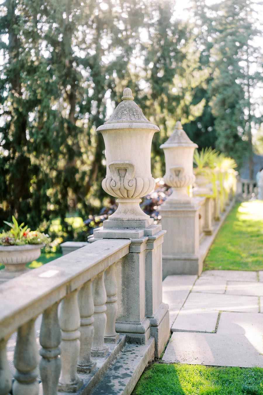 Elegant stone urns and balustrade in a sunlit garden, surrounded by lush greenery.