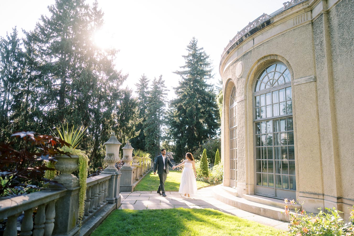 A couple holding hands walks toward a grand stone building in a lush garden setting with tall pine trees and bright sunlight filtering through, creating a romantic and picturesque scene.
