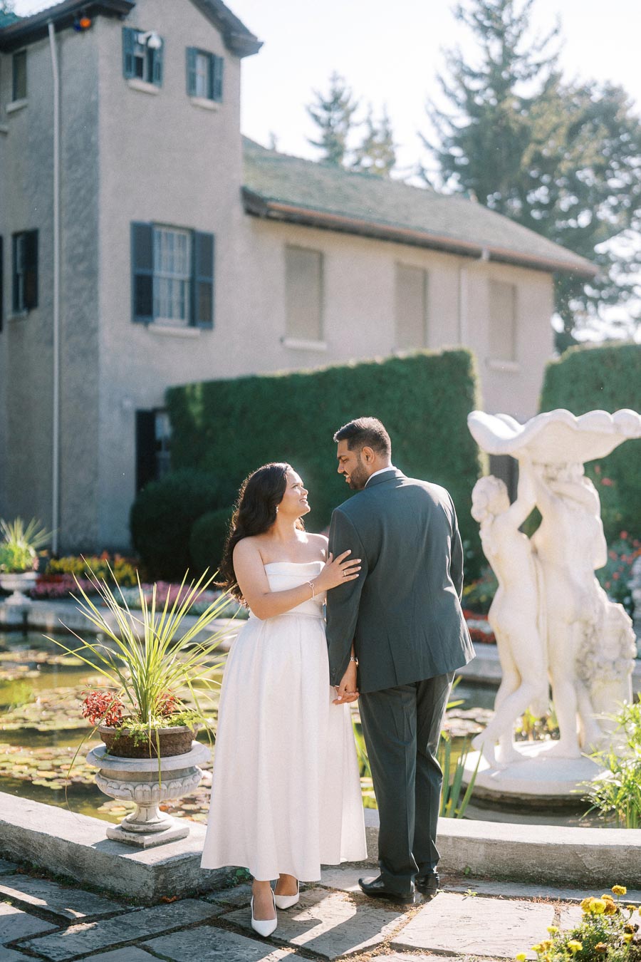 A couple sharing a romantic moment outdoors near a historic building and stone sculpture, surrounded by lush greenery and a tranquil pond.