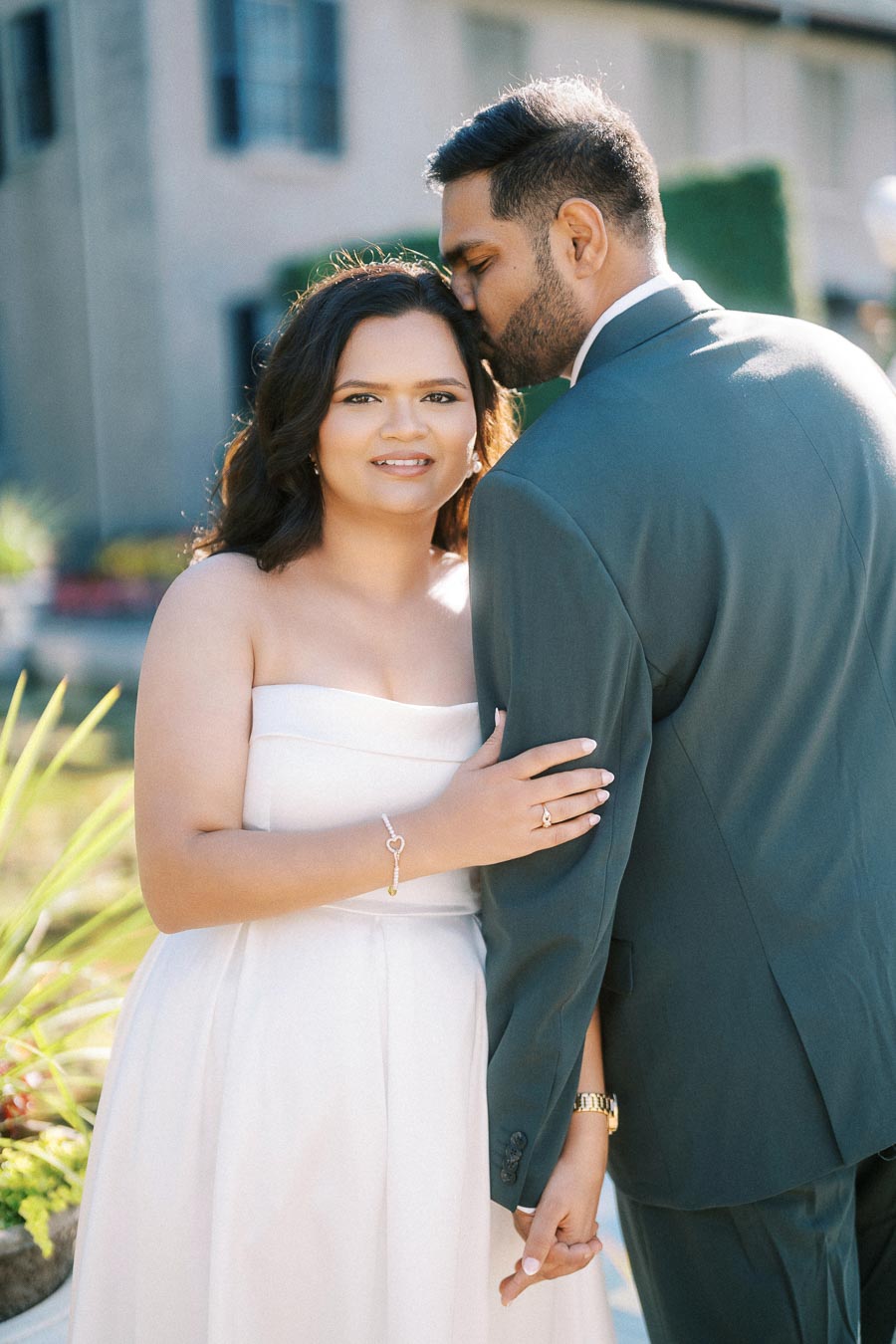 A couple embraces during an outdoor wedding photoshoot, with the bride in a strapless white dress and the groom in a dark suit, set against a garden backdrop.