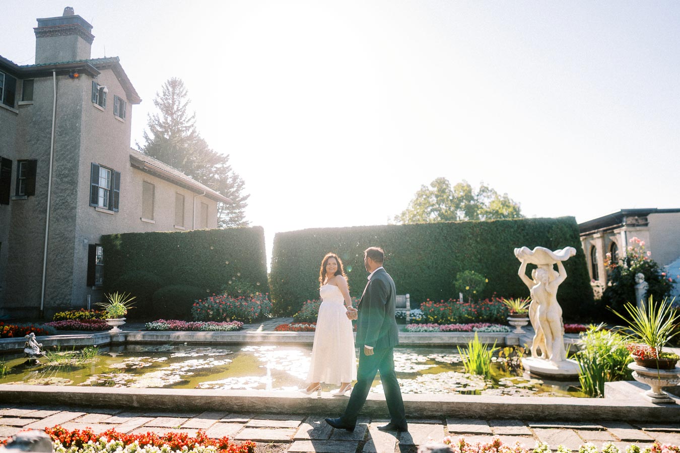 Couple enjoying a romantic stroll in a picturesque garden, surrounded by lush greenery, vibrant flowers, and classic statues, with a historic mansion in the background illuminated by bright sunlight.