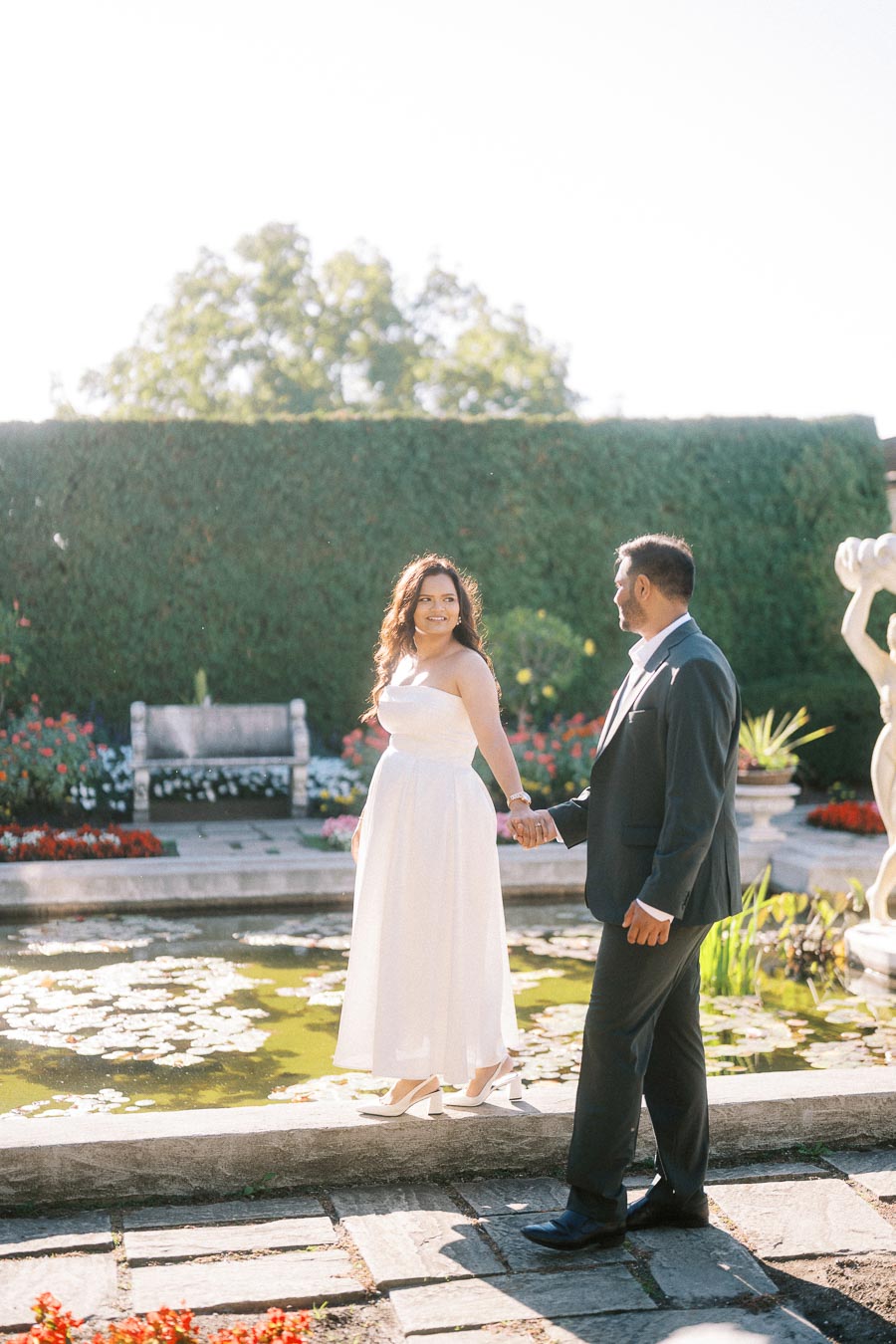 A couple holding hands and smiling in a lush garden setting, with a pond and flower beds surrounded by greenery.