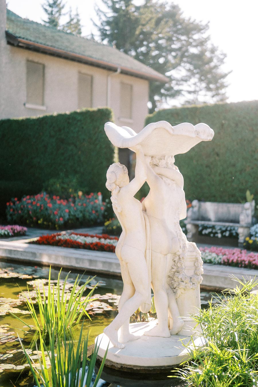 Elegant marble fountain sculpture in a garden setting, surrounded by vibrant flowers and greenery, with a house and neatly trimmed hedge in the background.