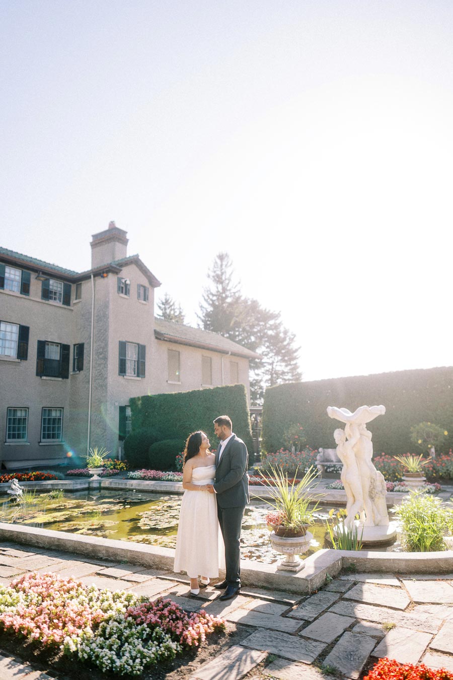 A couple in formal attire stands near a picturesque garden pond with a classical statue, surrounded by blooming flowers and a historic building in the background.