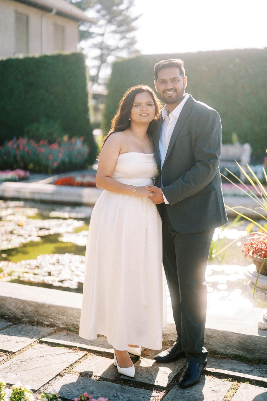 A couple standing together in a garden setting, with a pond and vibrant flowers in the background, dressed in formal attire.