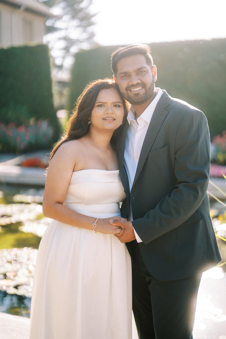 A couple in formal attire standing together in a garden setting with greenery and a pond in the background, smiling warmly at the camera.