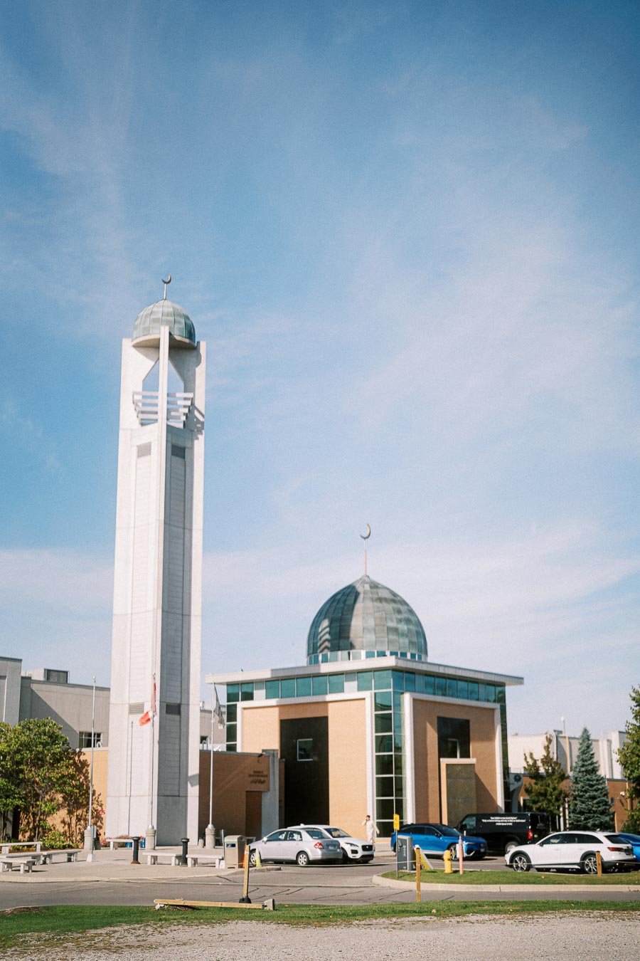 Modern mosque with a tall minaret and a domed roof under a clear blue sky, surrounded by parked cars.