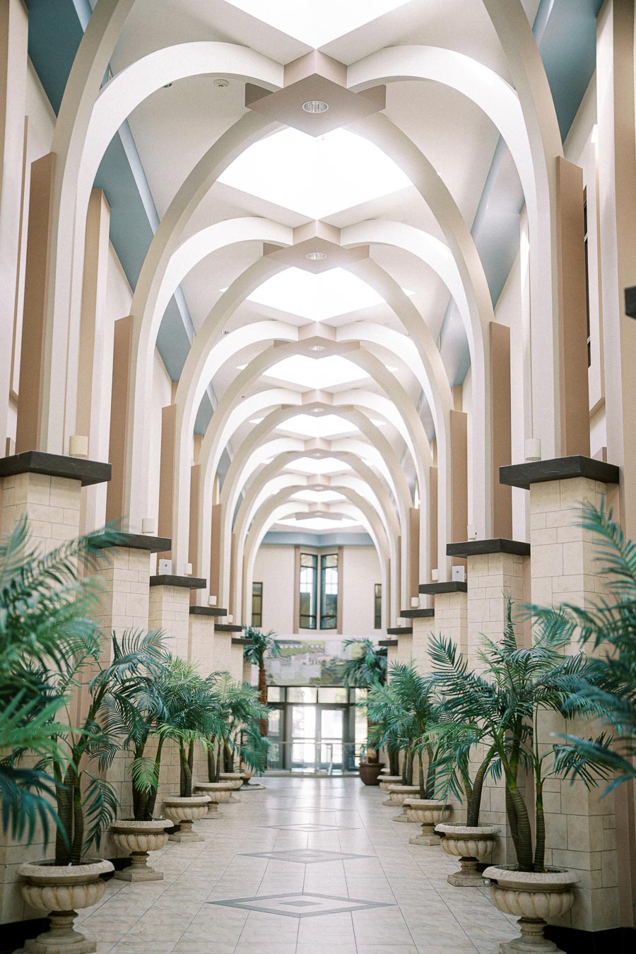 Elegant architectural interior of a hallway adorned with tall, arched ceilings and symmetrical rows of potted palm plants, featuring a polished tiled floor leading to a windowed exit.