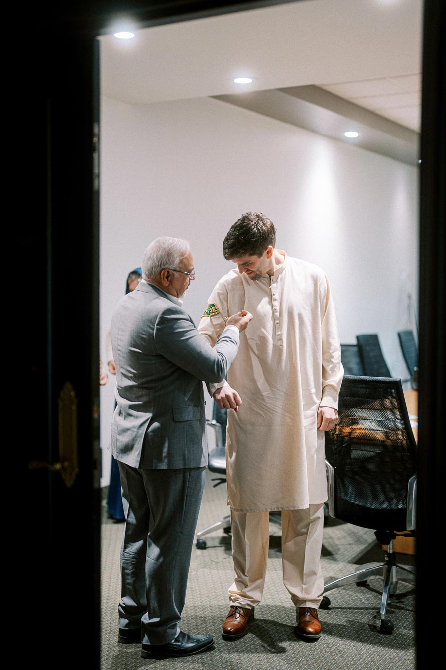 Two men in formal and cultural attire, one adjusting the other's sleeve, in a conference room setting.