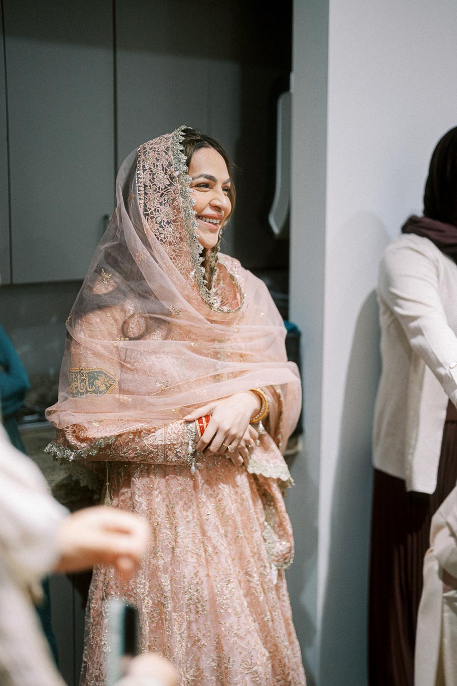 Smiling woman in a pink traditional dress with intricate embroidery and a matching dupatta, standing indoors.