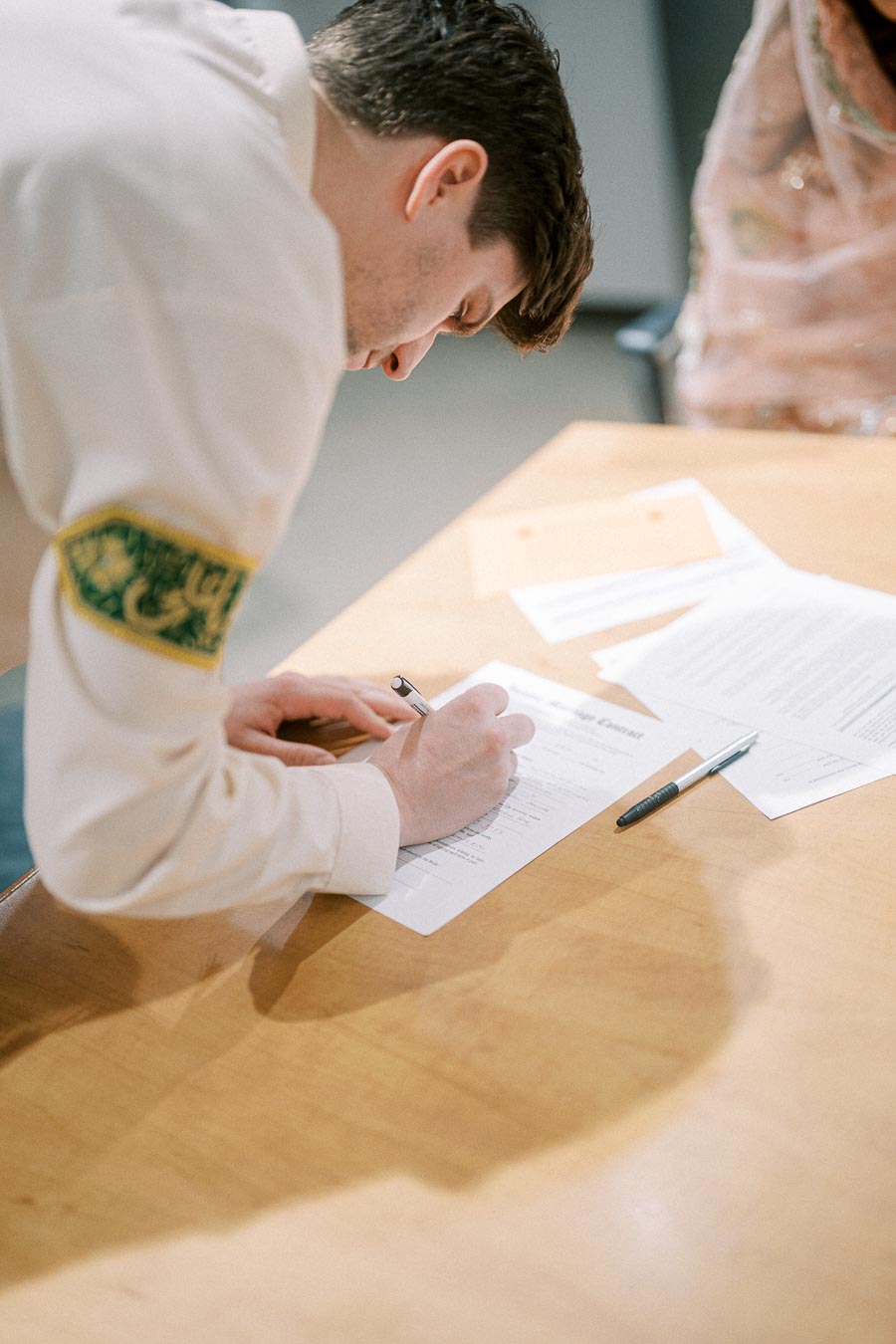 A person in a uniform signs a document at a wooden desk, with additional papers and pens nearby.
