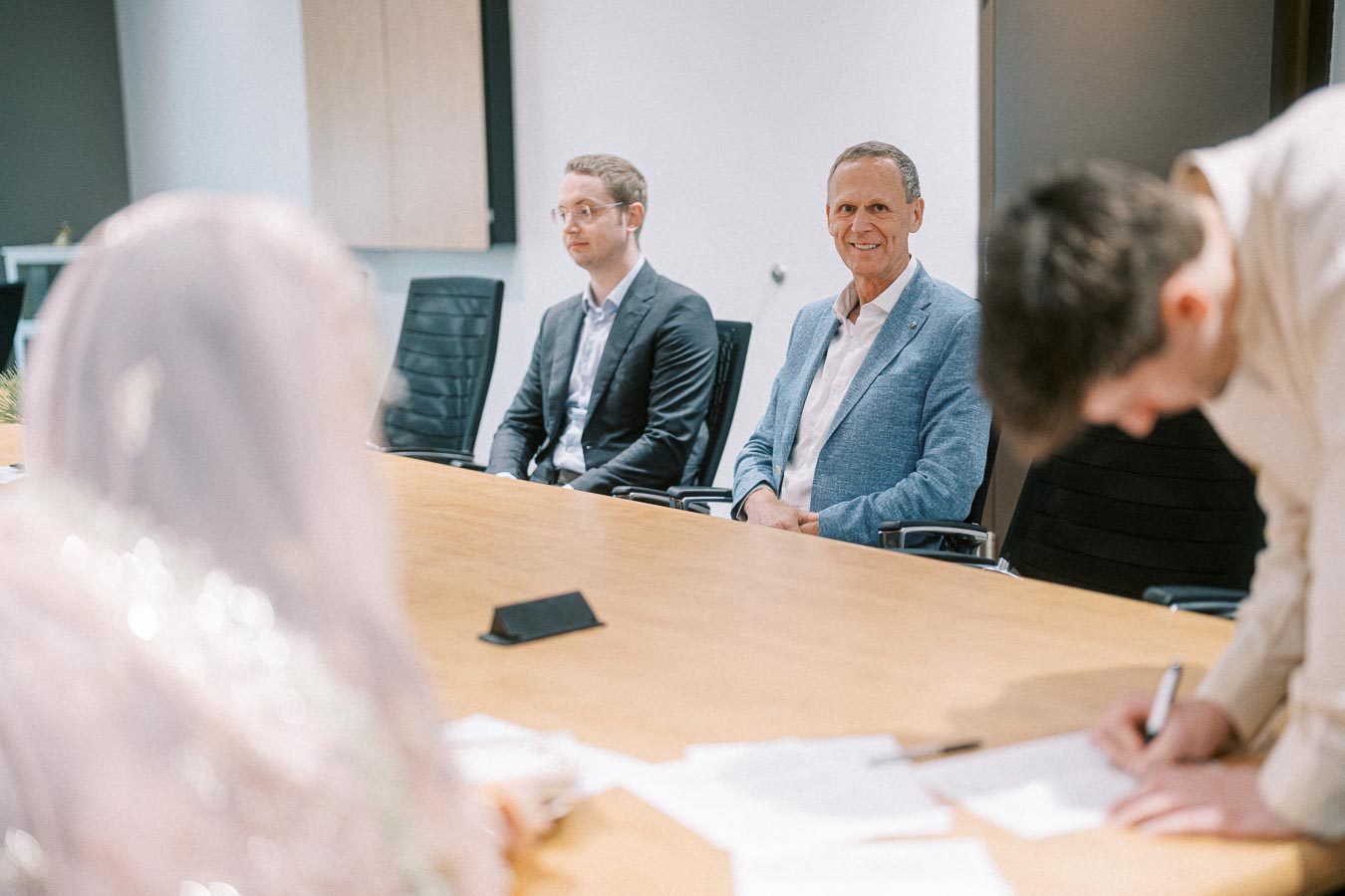 A professional meeting in a conference room with two men seated at the table, while another person signs documents.