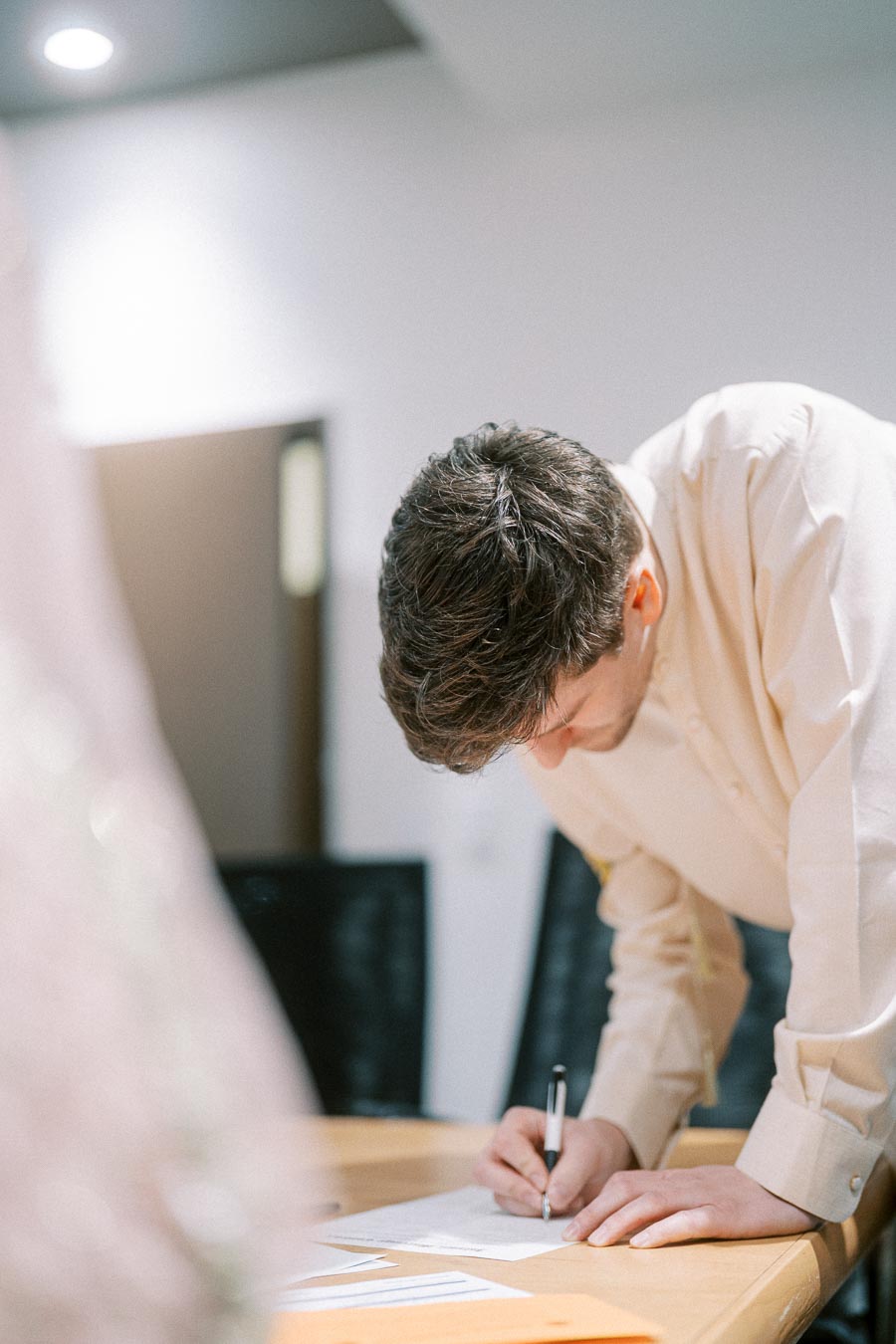 A person in a light-colored shirt signs a document on a wooden desk, with a focus on the signing process in an office setting.