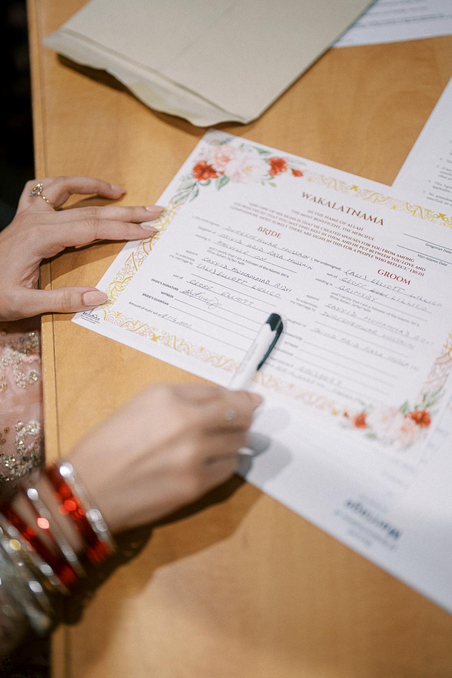 A person signing a marriage contract document on a wooden table, showcasing cultural wedding traditions with intricate floral designs on the certificate.