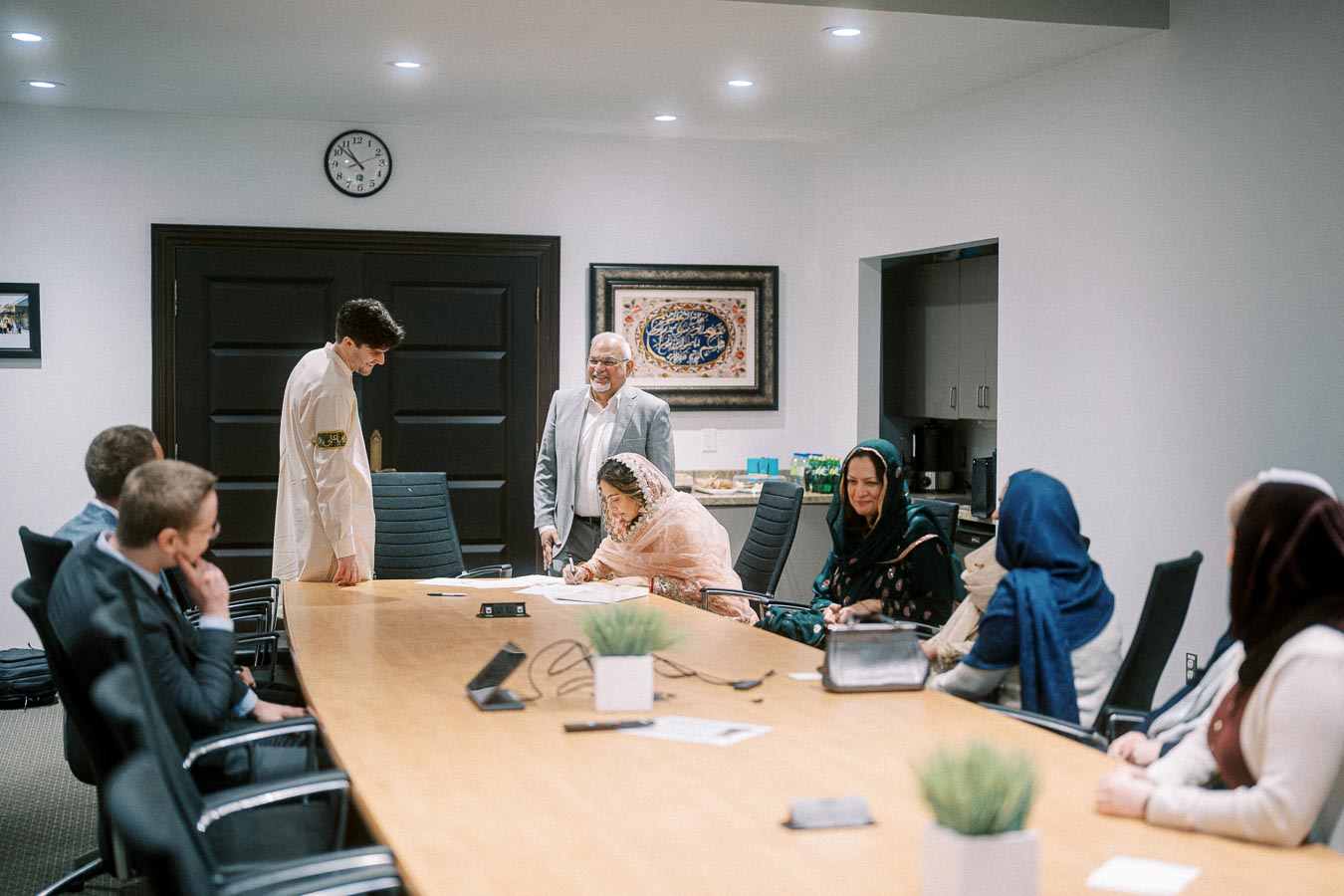 A group of people gathered around a conference table in a meeting room, with one woman signing a document while others look on.