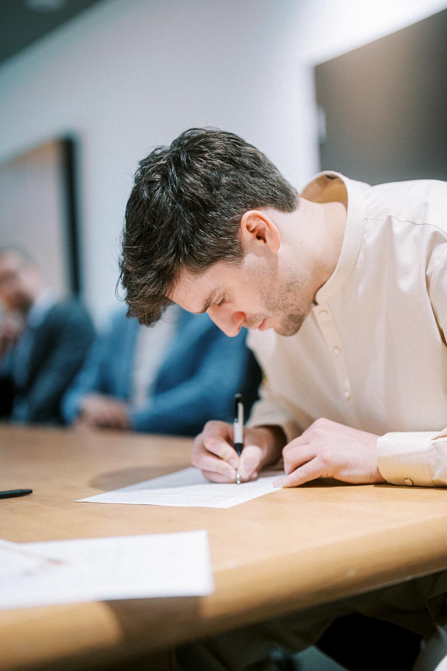 A man in a white shirt signs a document at a wooden table, with blurred people in the background, in an office setting.