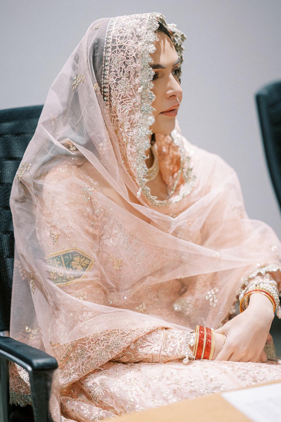A person wearing a traditional peach-colored outfit adorned with intricate lace and embroidery, sitting in a chair. The attire includes a veil with detailed patterns and sparkling embellishments, and the individual is wearing red and gold bangles.