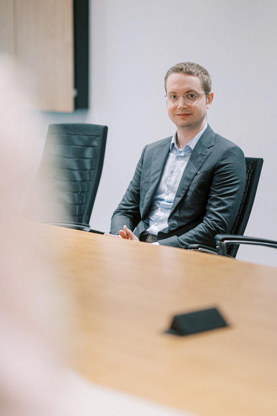 Man in a suit sitting in a conference room chair, attending a meeting at a wooden table.