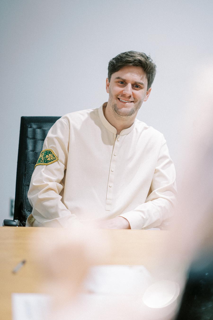 Smiling man in a white shirt with decorative patch, sitting at a desk in a modern office setting.