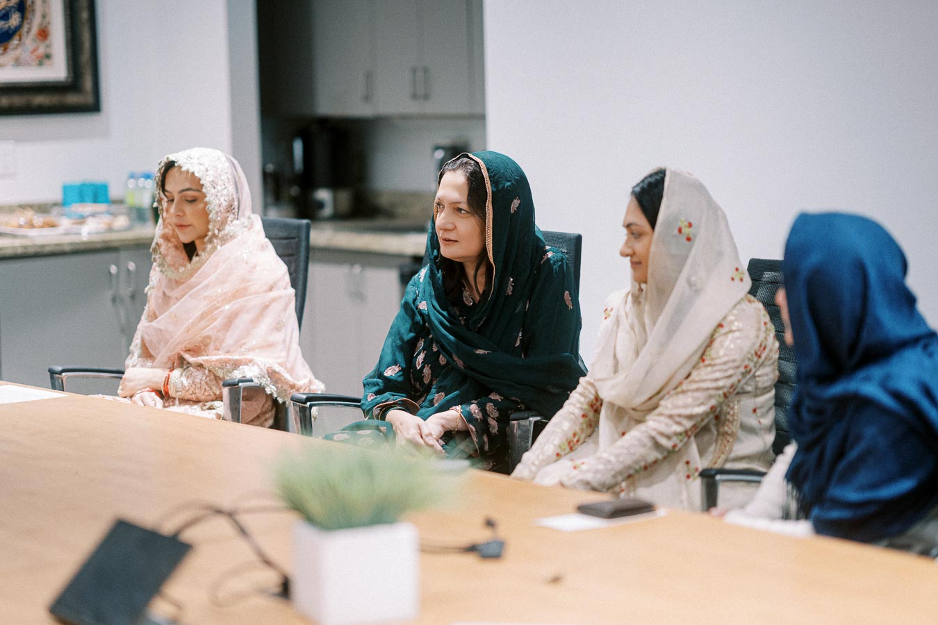 Four women wearing traditional cultural attire sit in a conference room, engaged in conversation. The room has modern office furniture and decor.