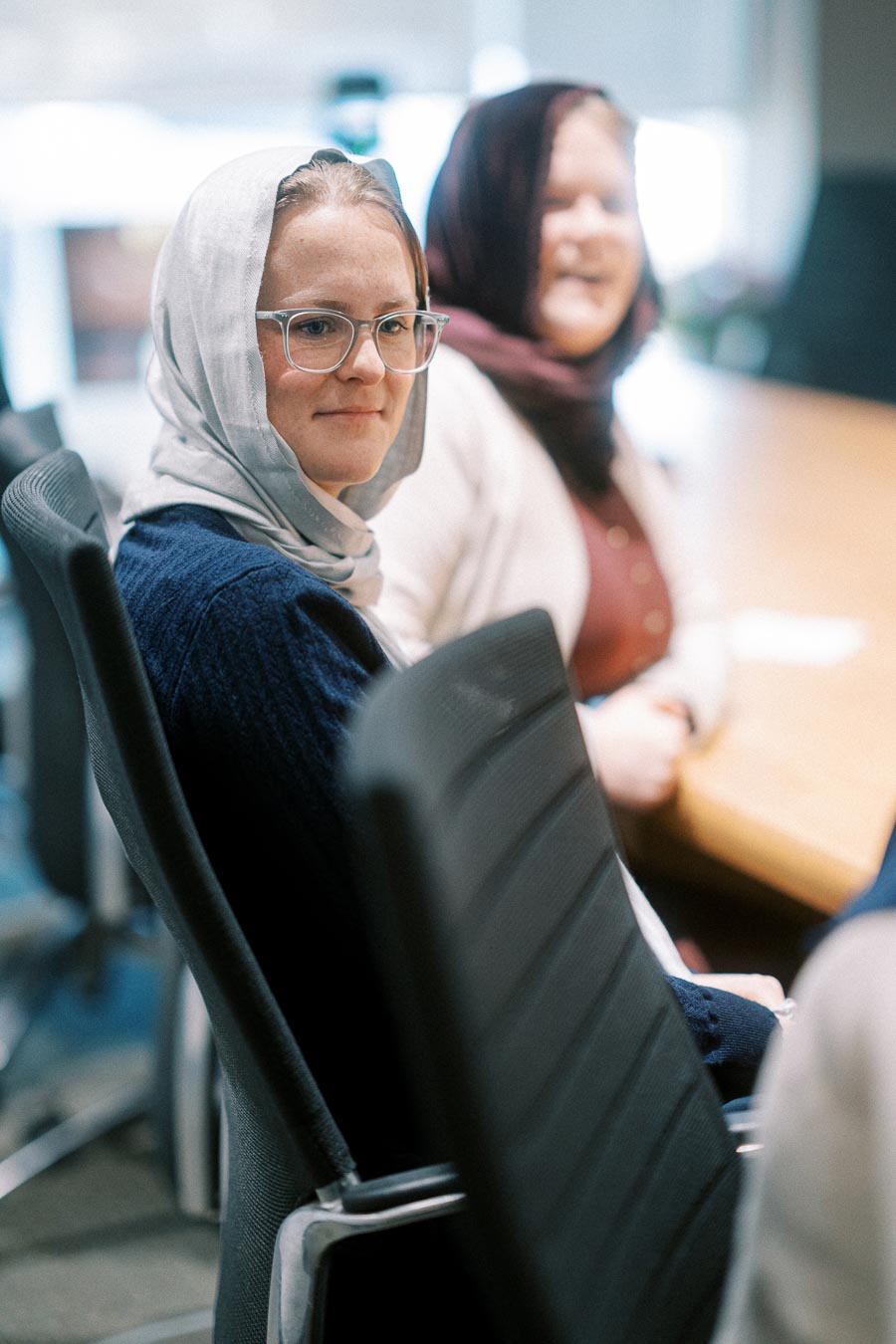 Two women wearing headscarves seated in office chairs during a meeting, one smiling and the other looking attentive.