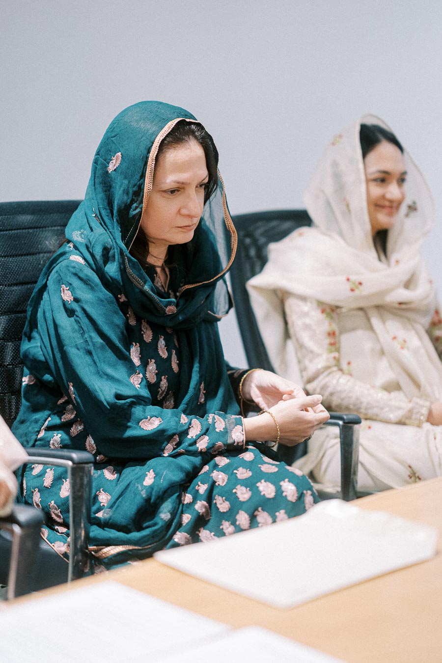 Two women in traditional attire sitting in an office meeting room, focused and engaged, wearing embroidered shawls.
