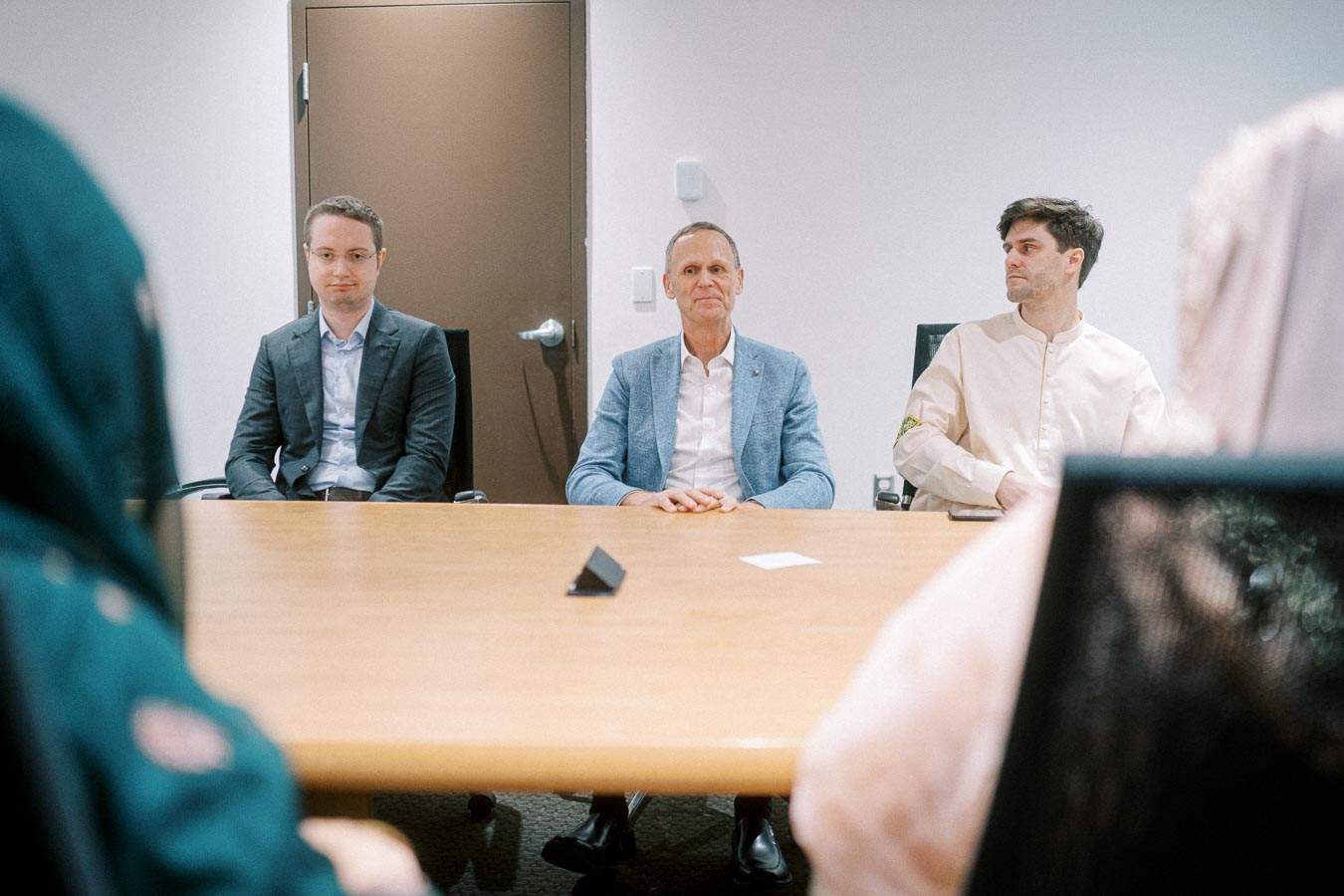 A group of business professionals sitting around a conference table during a meeting, with two men in formal attire and others partially visible, focusing on collaboration and discussion in a modern office setting.