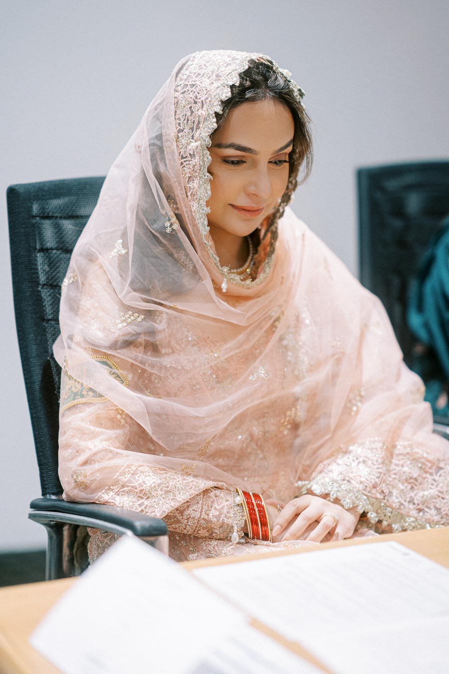 A woman in a traditional pink embroidered outfit and headscarf, sitting at a desk in a formal setting.