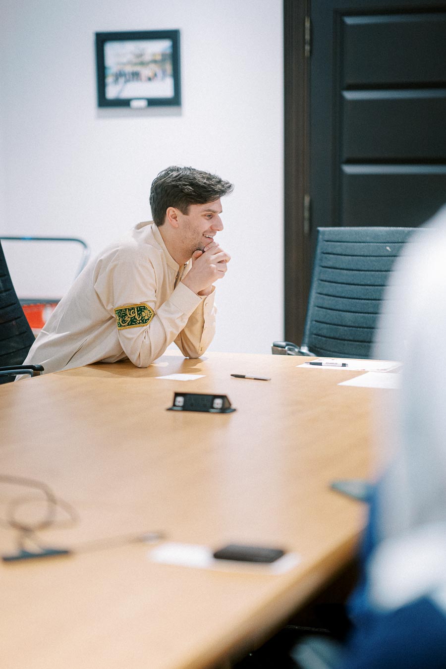 Man sitting at a conference table, wearing a traditional long-sleeve garment, focused and thoughtful during a meeting in a modern office setting.