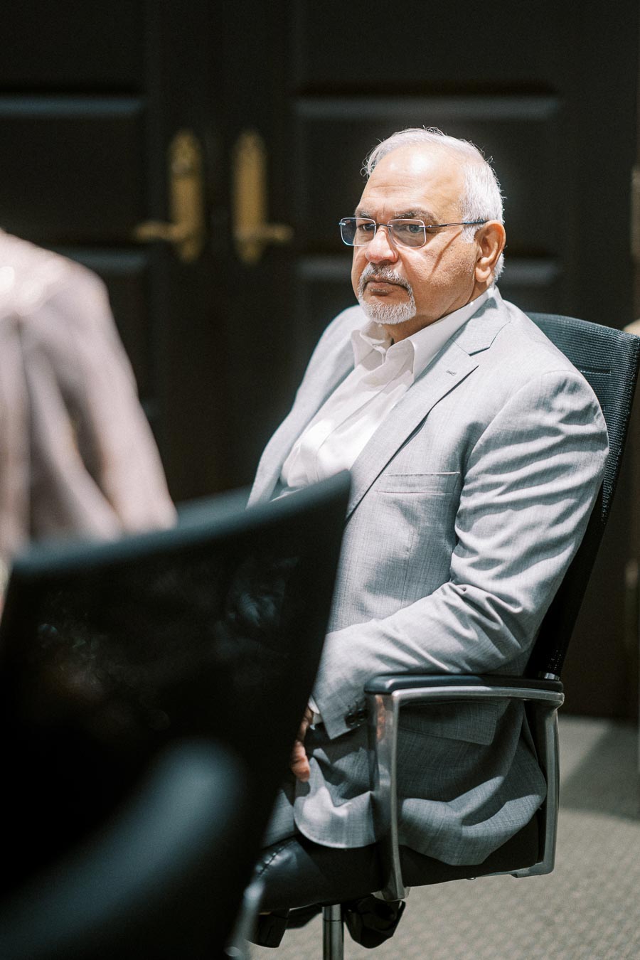 A man in a business suit sits attentively in a conference room, looking thoughtfully ahead, suggesting a professional meeting or corporate discussion environment.