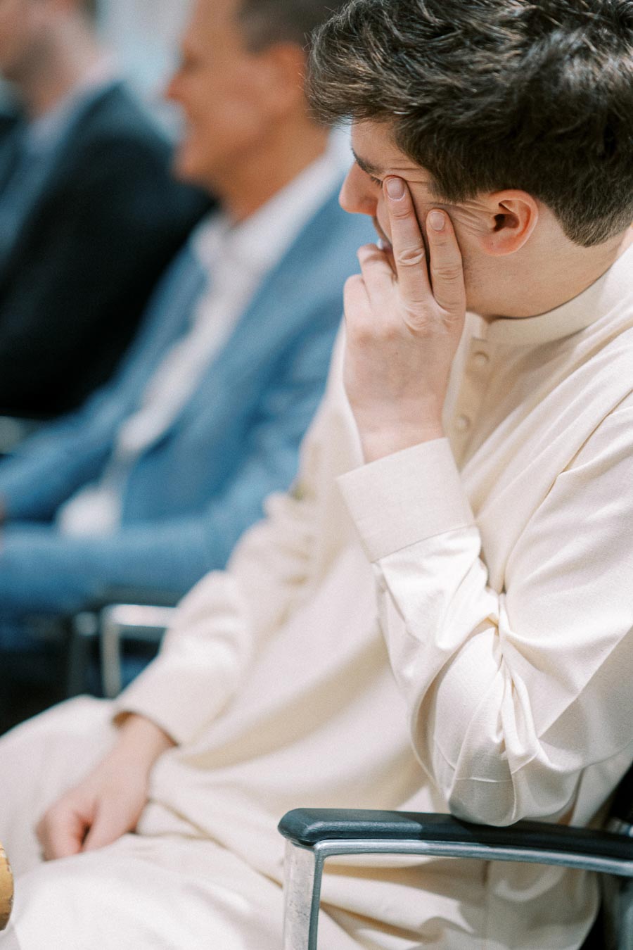 A person sitting in a light-colored traditional outfit at a conference or meeting, resting their head on their hand, with blurred people in the background.