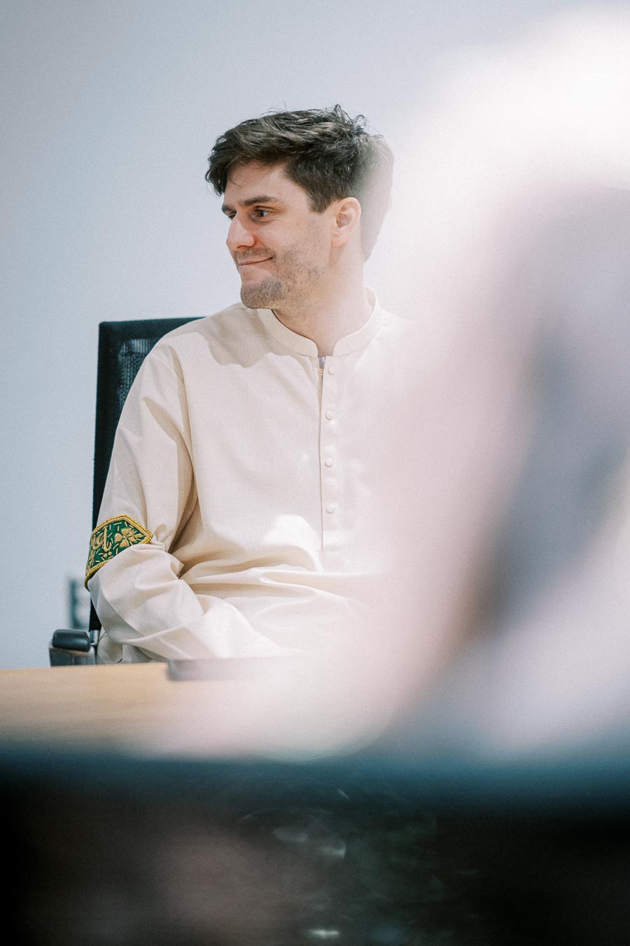A person in a light-colored traditional outfit sits in an office chair, smiling and looking to the side, with soft lighting creating a serene atmosphere.