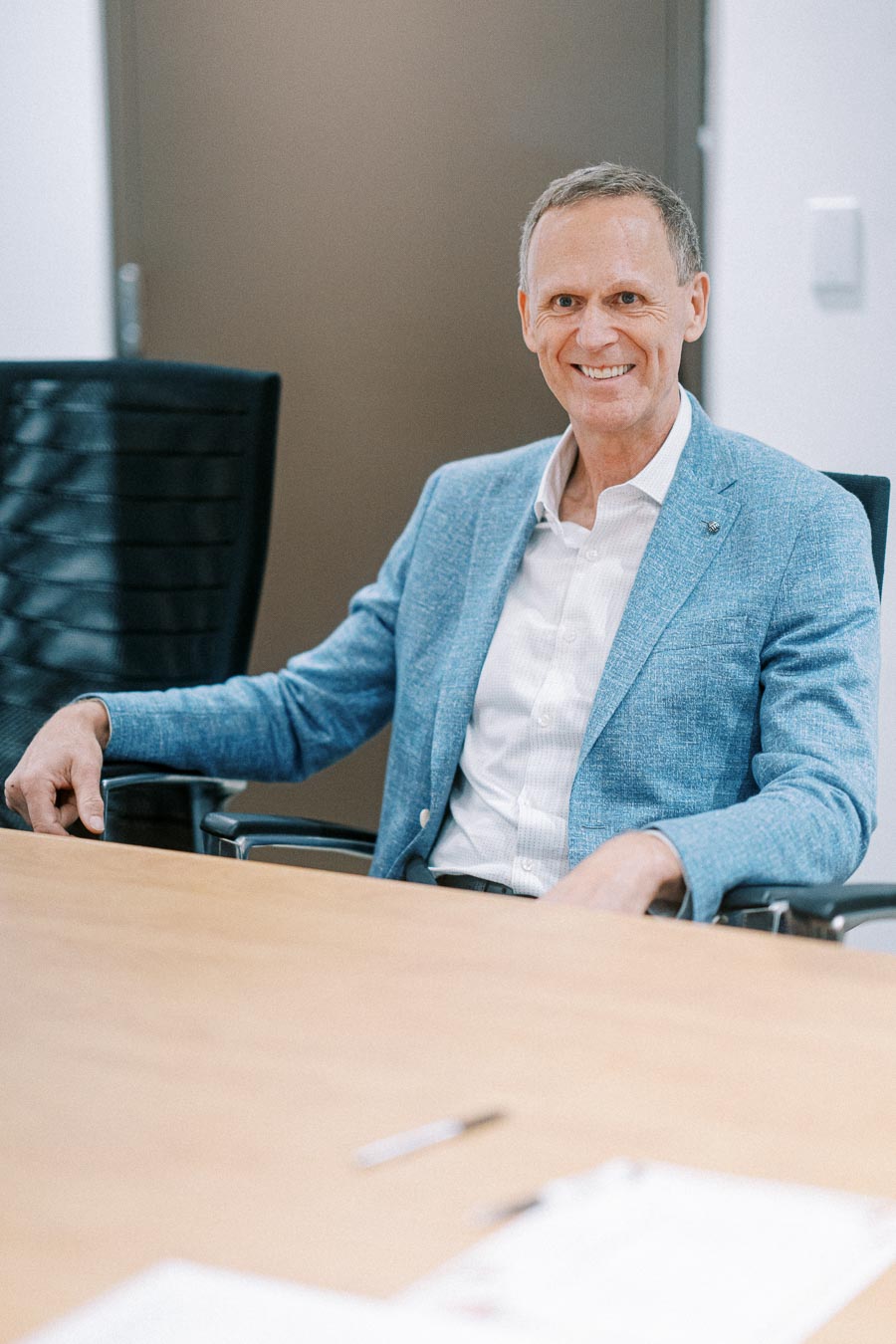 A man in a blue blazer sitting at a conference table, smiling confidently in an office setting.