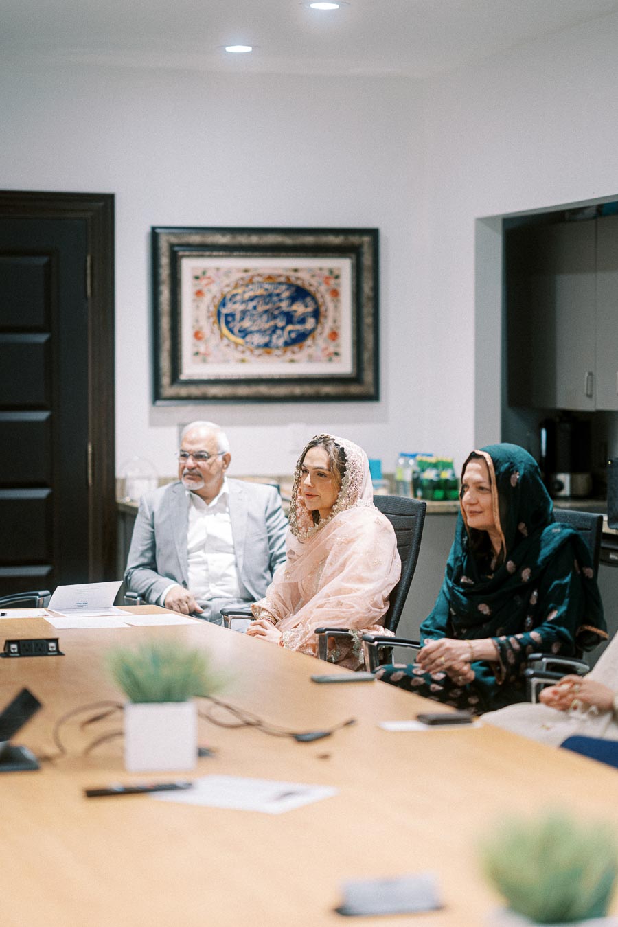 Three people in traditional attire sitting at a conference table in a modern meeting room, with a framed artwork on the wall in the background.