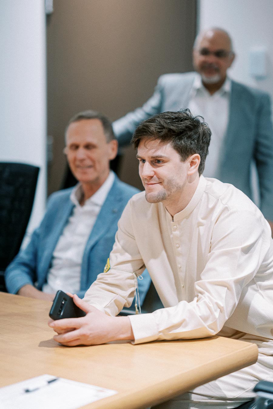 Group of professionals engaged in a business meeting, focused young man in a light-colored shirt holding a smartphone, with colleagues in suits in the background.
