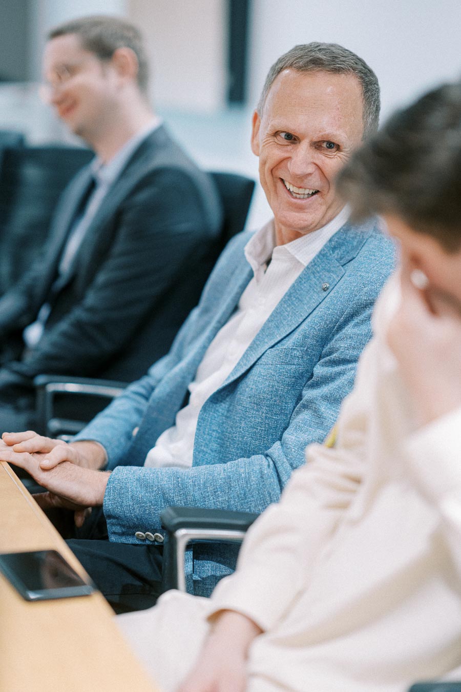 Professional business meeting with a smiling man in a blue blazer, engaged in conversation with colleagues, seated around a conference table.