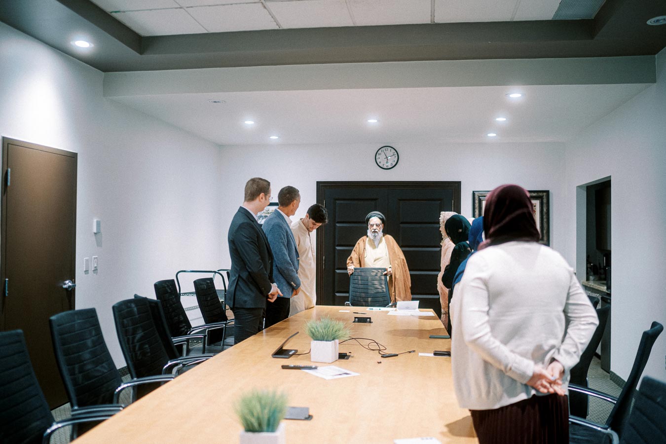 A group of diverse individuals standing around a conference table in a modern meeting room, engaged in a discussion with an elder in traditional attire at the head of the table, under a wall clock.