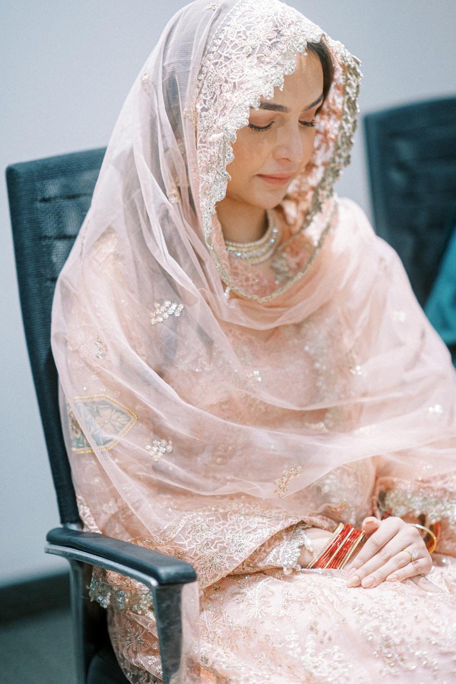 Young woman in a beautifully embroidered peach traditional outfit with a delicate lace veil, seated with downcast eyes and hands clasped, possibly in a reflective or prayerful moment, inside a modern room.