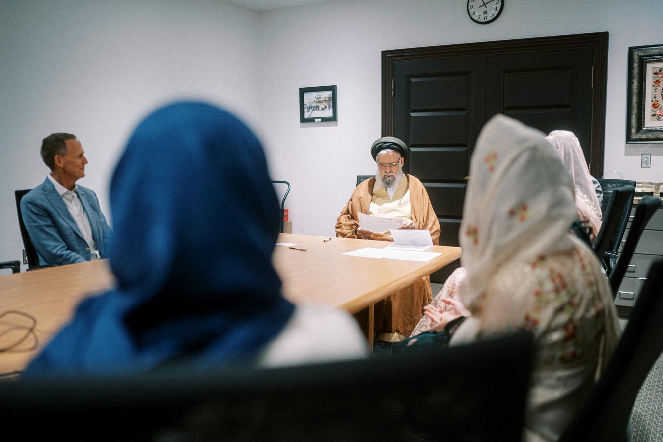 A group meeting in a conference room with a diverse group of people, including a person wearing traditional attire and others in business suits, having a discussion around a wooden table.