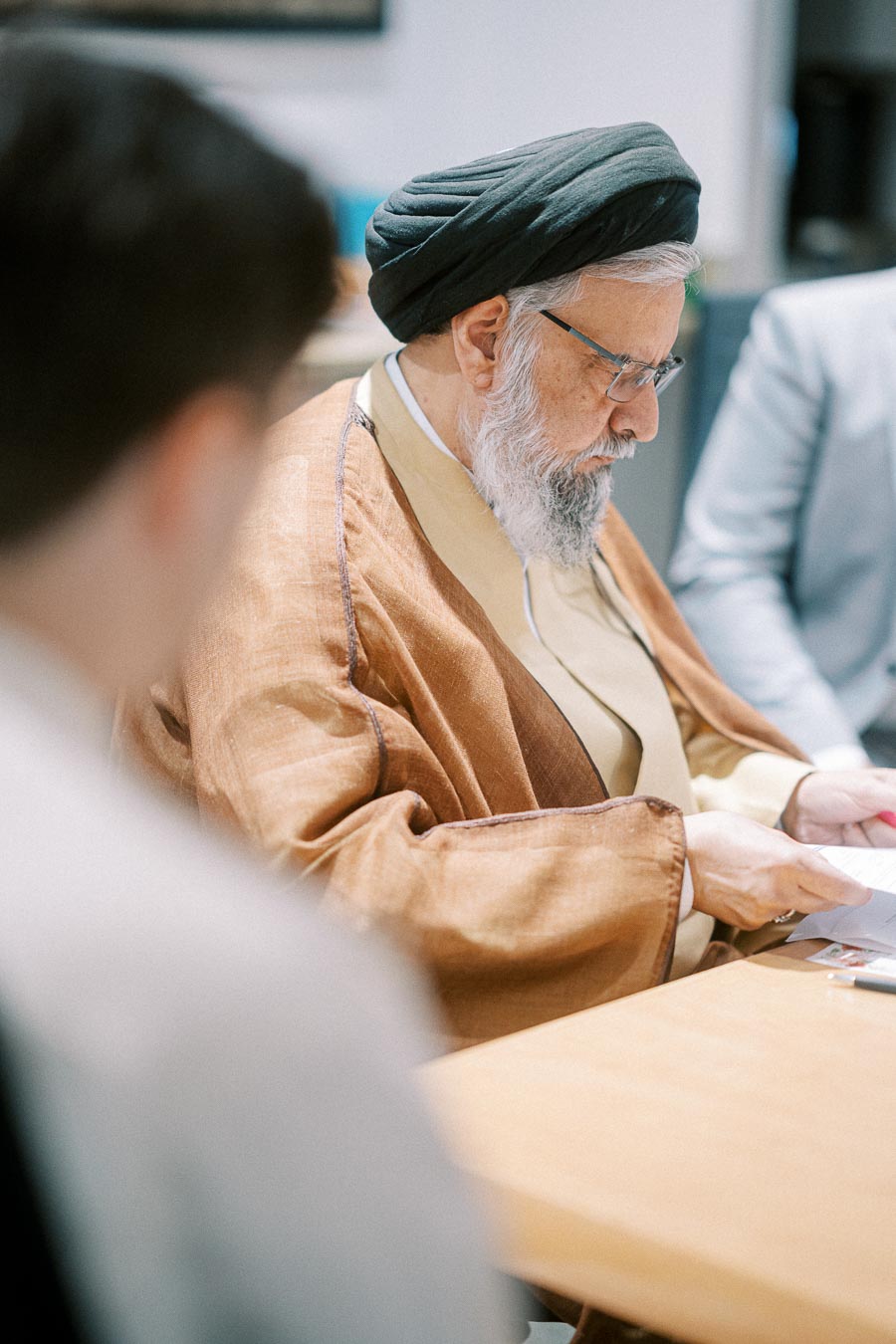 A bearded man in traditional attire, wearing a black turban and glasses, sits at a table reading a document.