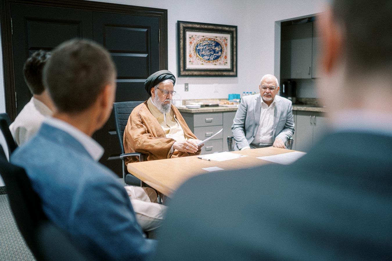 Meeting in a modern office setting with diverse individuals around a conference table, featuring a framed artwork on the wall and refreshments on a side counter.