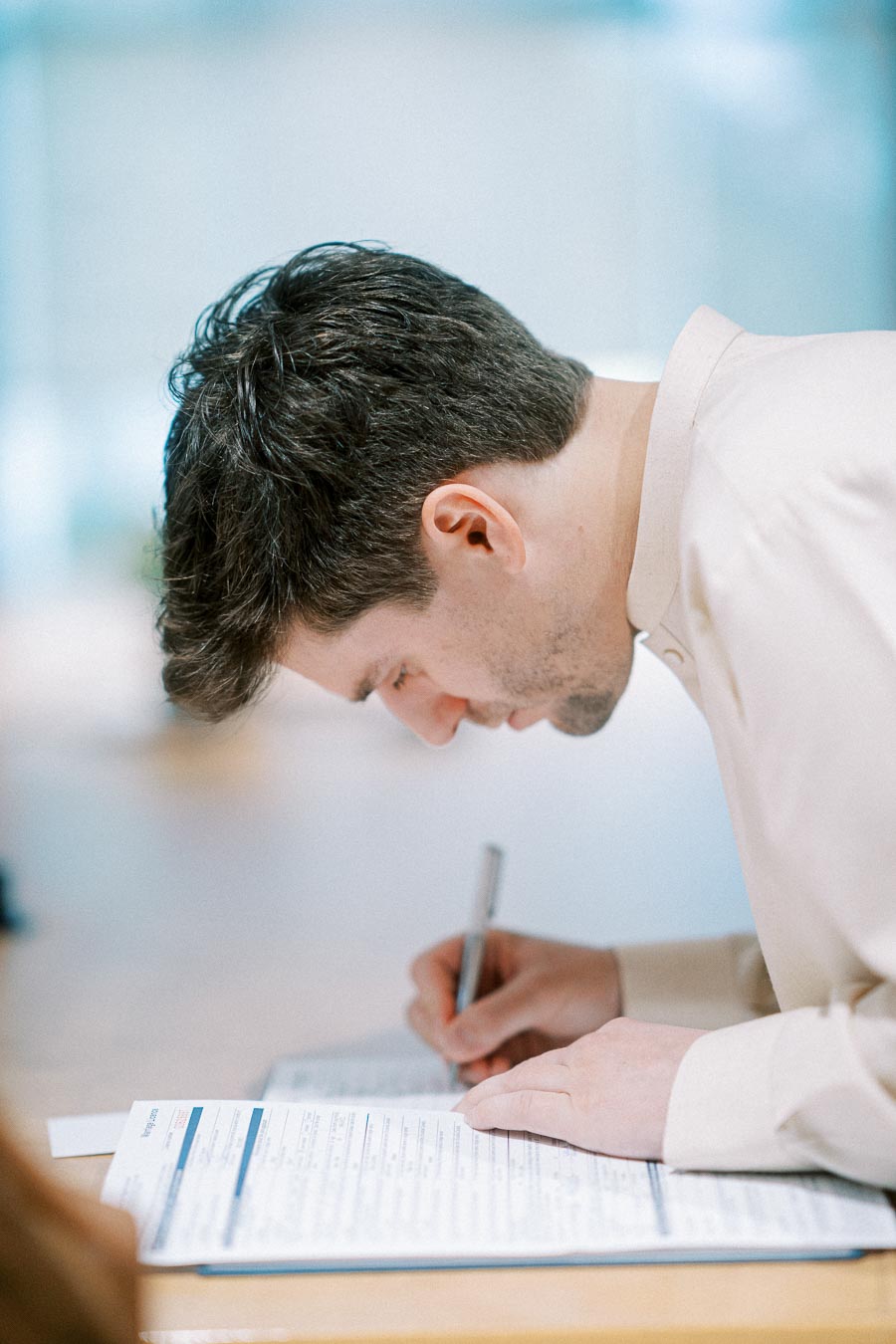 A person in a beige shirt writing on a document with a pen at a table, focusing intently.