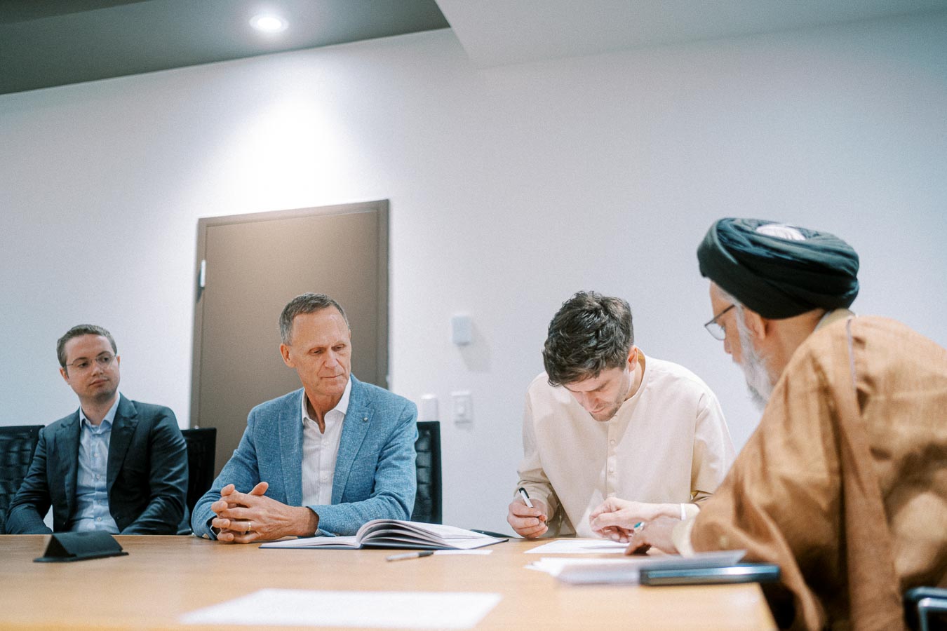 Four men in a business meeting are seated around a conference table. One man is signing a document while the others observe attentively. The setting appears professional, with papers and notebooks on the table.
